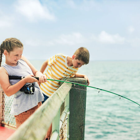 Two children are fishing from a pier, with one girl holding a fishing rod and a boy leaning over the railing looking down at the water. The background features a calm ocean under a partly cloudy sky.