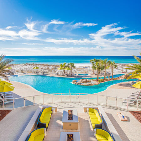 A beachside resort pool area with lounge seating, palm trees, and a view of the ocean under a clear blue sky.