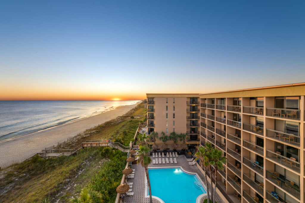 Aerial view of a beachfront hotel with a swimming pool, adjacent to a sandy beach and ocean at sunset.