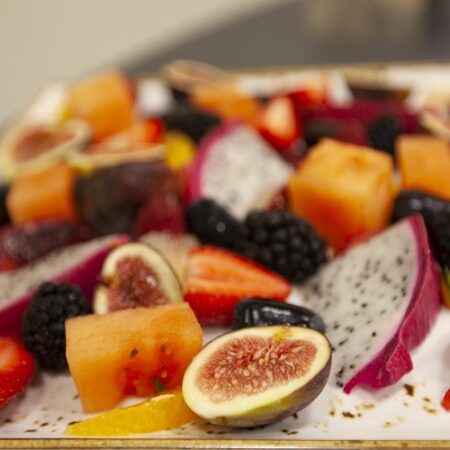 A close-up of a colorful fruit platter featuring sliced strawberries, figs, blackberries, dragon fruit, watermelon, and oranges arranged on a white rectangular plate.