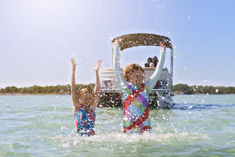 Two kids play in the calm waters of Destin-Fort Walton Beach, Florida with their parents nearby on a boat. 