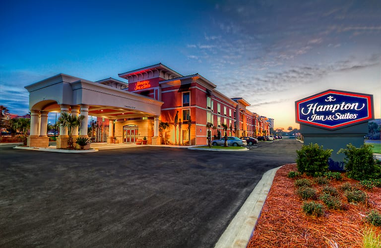 Exterior view of a Hampton Inn & Suites hotel at dusk, with a lit sign in the foreground and cars parked near the entrance.