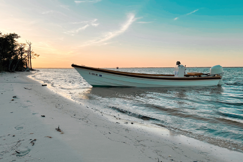 A small boat is moored on a sandy beach at sunset, with calm sea and sky showing gradient colors of blue and orange.
