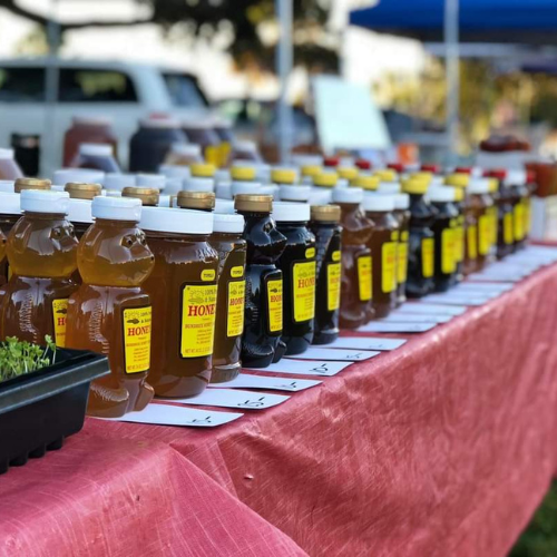 Row of honey bottles on a table at a farmers market, labels facing forward, under natural light with blurred background.