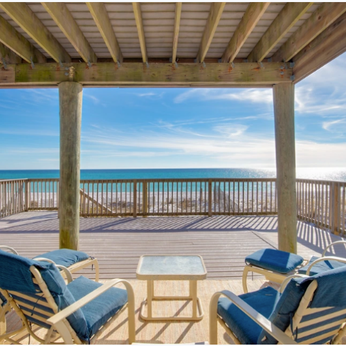 Deck with four padded lounge chairs and a small table under a wooden pergola, overlooking a clear blue ocean and bright sky.