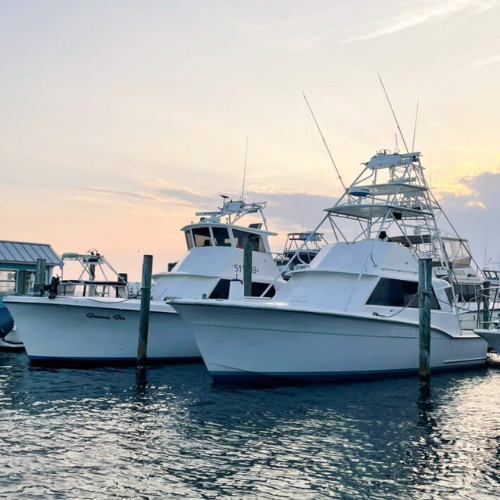 Two white boats are docked at a marina during sunset. The water is calm, and the sky shows hues of orange and blue.