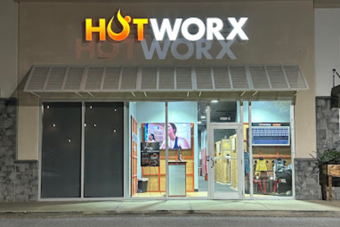 Front entrance of a Hotworx fitness studio at night, illuminated sign above the door, and interior visible through large windows.