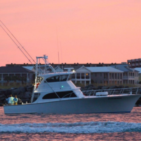 A fishing boat with multiple fishing rods is on the water near the shore at sunset, with buildings visible in the background.