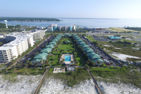 Aerial view of a coastal resort featuring multiple buildings with green roofs, a swimming pool in the center, and a boardwalk leading to the beach. Surrounding areas include parking and nearby waters.