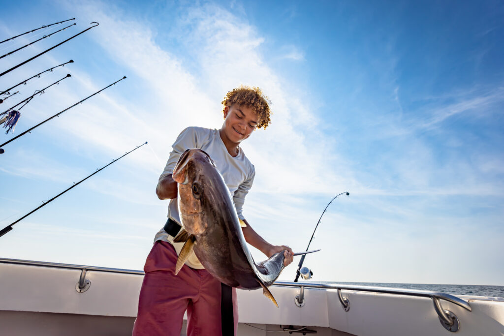 A person on a boat holds a large fish with fishing rods in the background under a clear, blue sky.