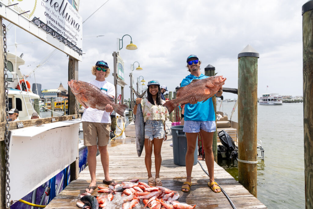 Three people stand on a dock holding large fish, with a pile of caught fish at their feet and boats docked in the background.