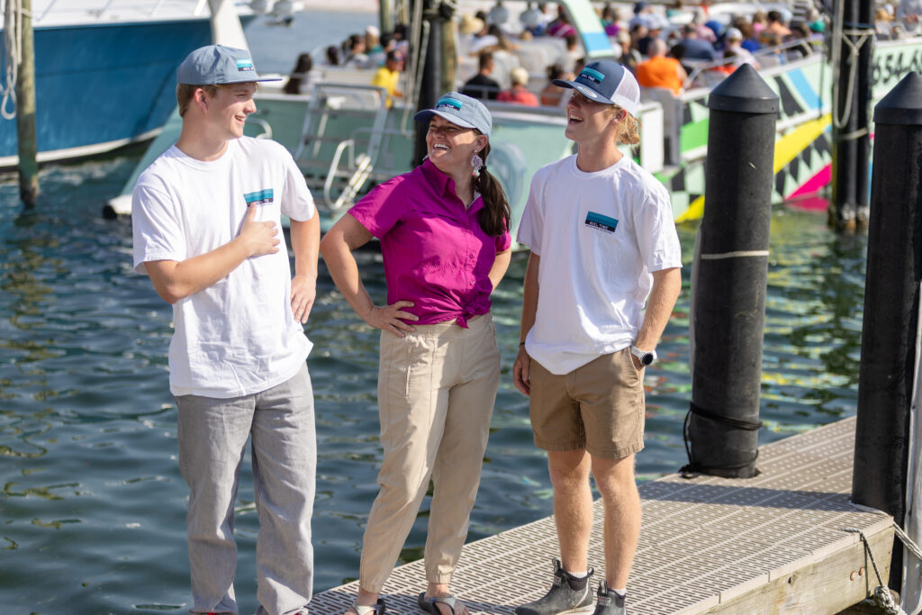 Three people stand on a dock near boats, wearing casual clothes and caps, smiling and talking on a sunny day.