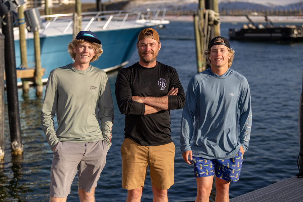 Three men stand on a dock by the water, boats in the background. They are smiling and dressed in casual, sun-protective clothing suitable for boating or fishing.