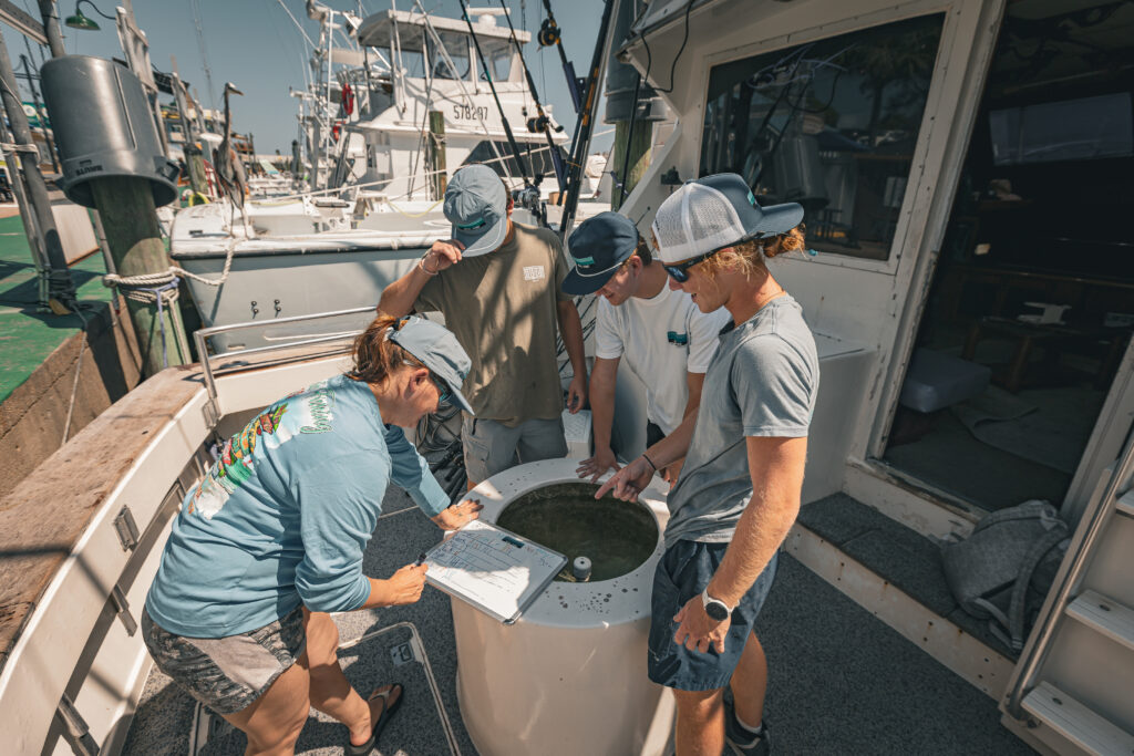 Four people on a fishing boat examine a circular tank, with fishing rods and other boats visible in the background.