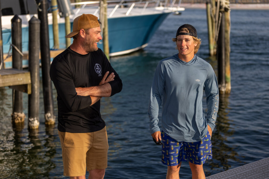 Two men stand on a dock by the water, talking and smiling. A boat is docked behind them. Both are wearing casual clothes suitable for warm weather.
