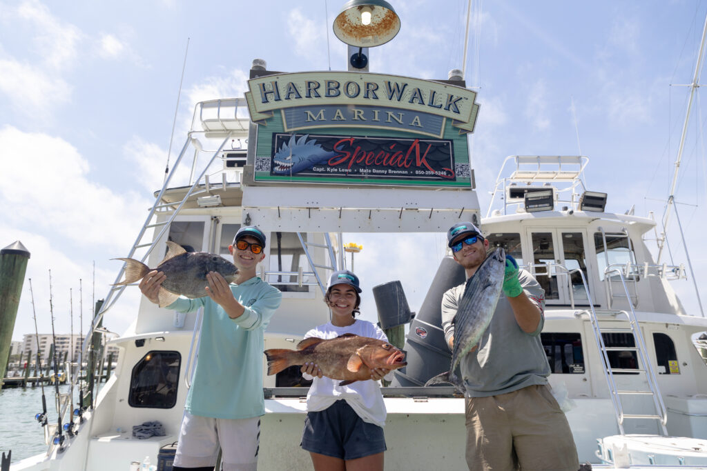 Three people stand in front of a boat at Harborwalk Marina, each holding a large fish they caught.