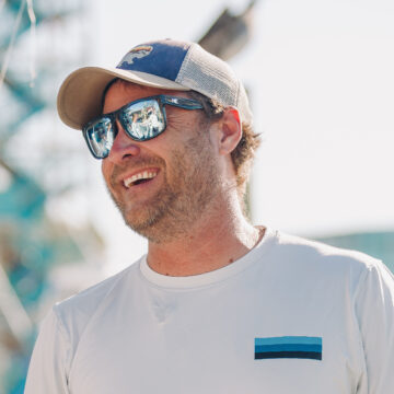 Man wearing a cap and reflective sunglasses smiles outdoors, with a blurred background of structures.