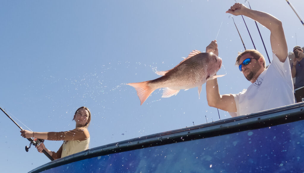 Two people fishing on a boat, with one person holding up a large fish. The other person is casting a line into the water. The sky is clear and blue.