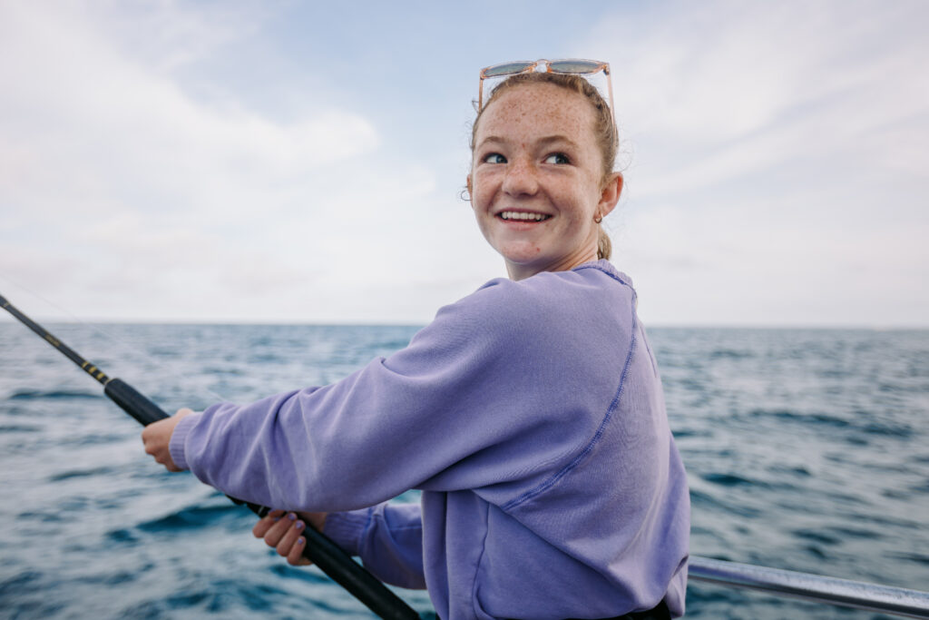 A person in a purple sweatshirt smiles while holding a fishing rod on a boat in the open ocean.