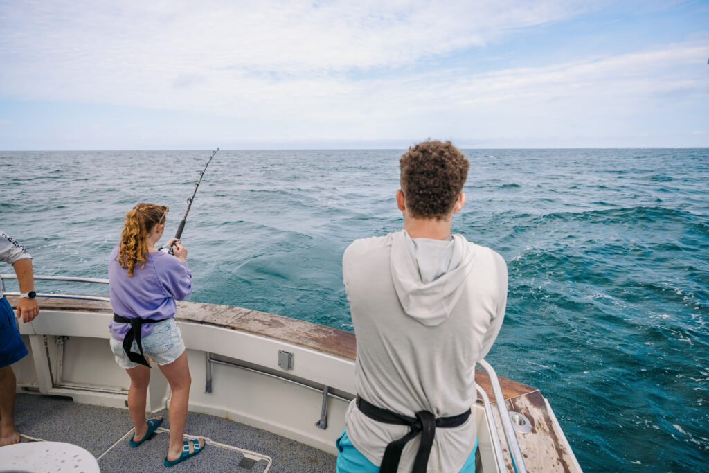 Two people stand on a boat deck facing the ocean; one is fishing with a rod, and the other watches.