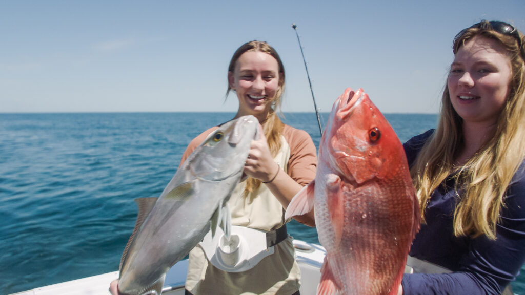 Two people on a boat, each holding a large fish against the backdrop of a clear blue ocean and sky. One fish is silver, and the other is red.