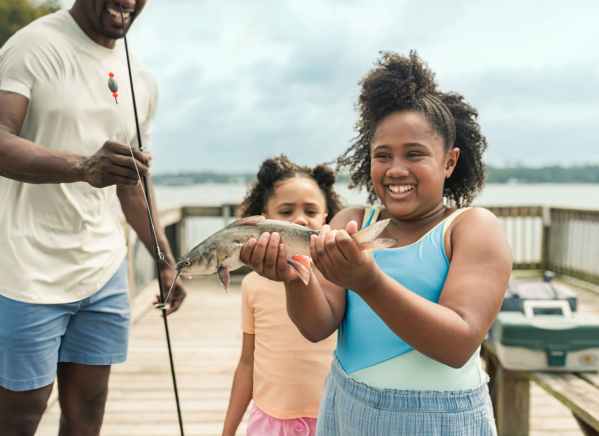 A smiling girl holds a fish while standing on a pier with a younger girl and a man holding a fishing rod nearby.