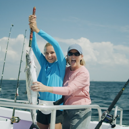 Three people on a boat holding up a large fish with fishing rods in the background on a sunny day.
