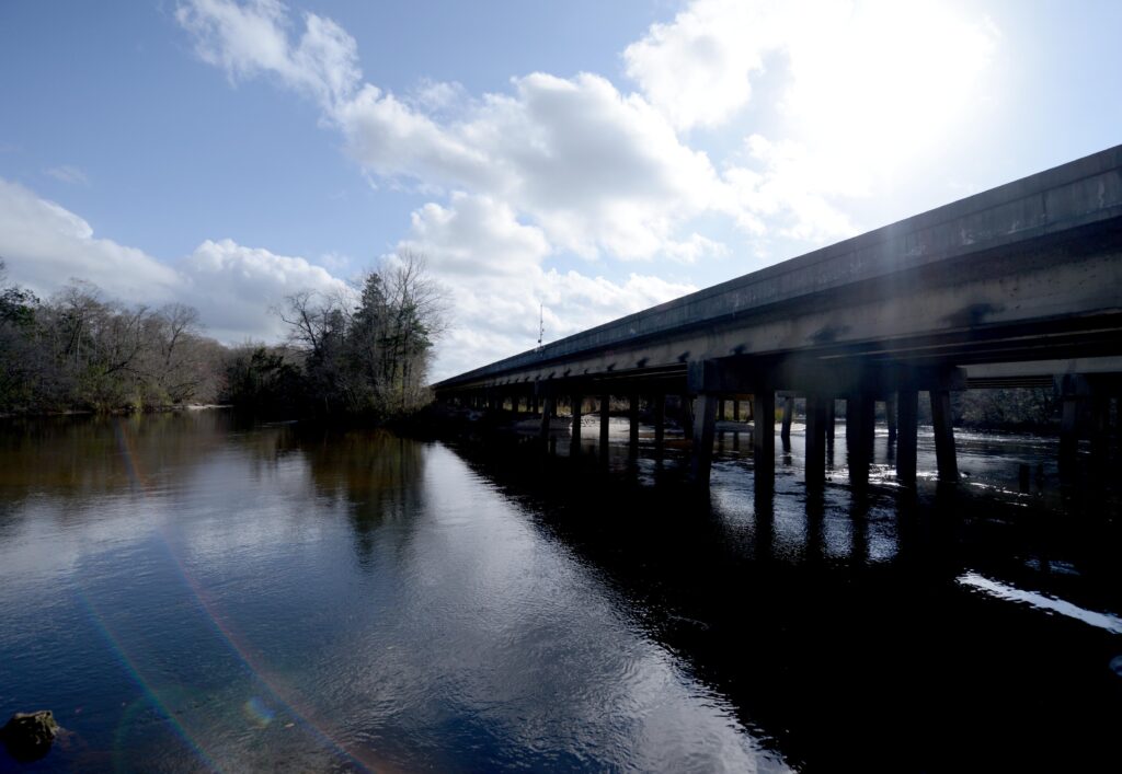 A long bridge spans across a calm river with a clear blue sky and scattered clouds overhead. Trees line the riverbank on the left side.