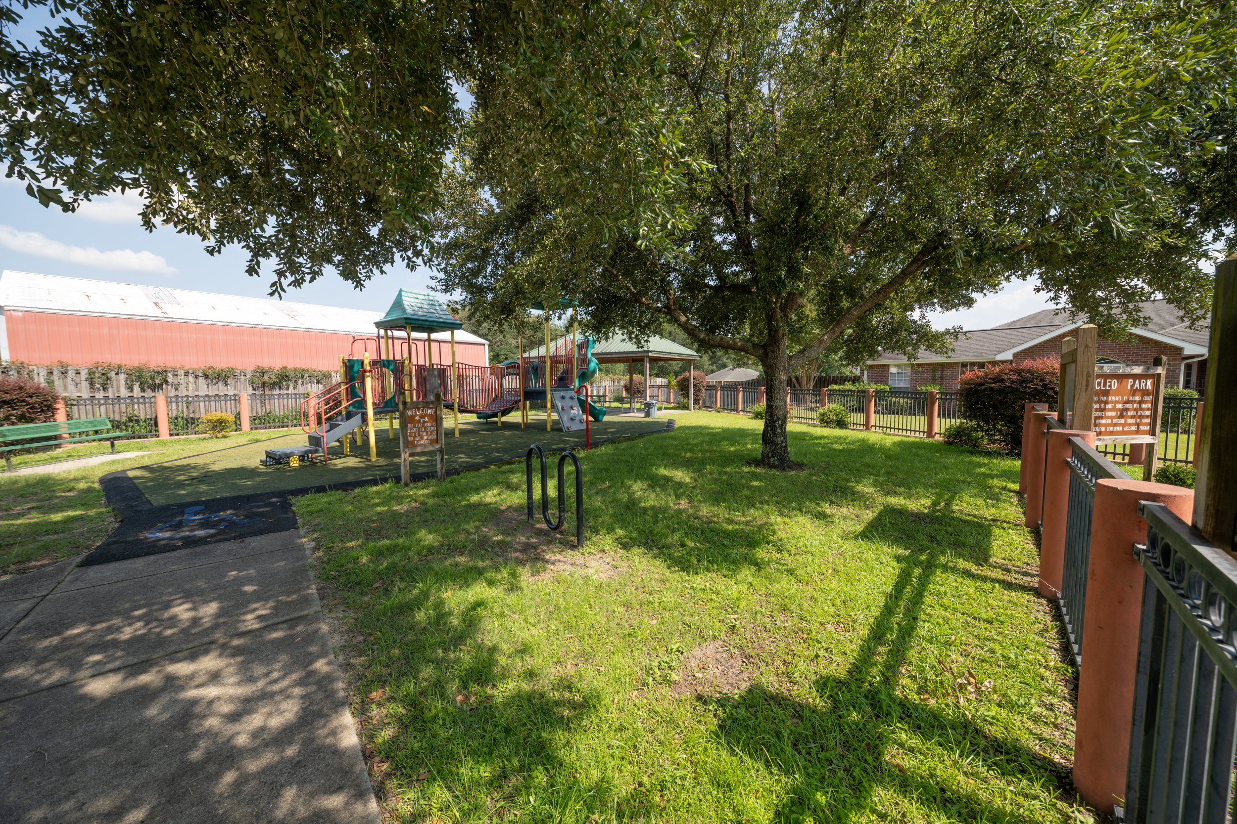 A fenced playground with slides, climbing structures, and a bench shaded by trees. A paved walkway runs through the grass, and a gazebo is visible in the background.