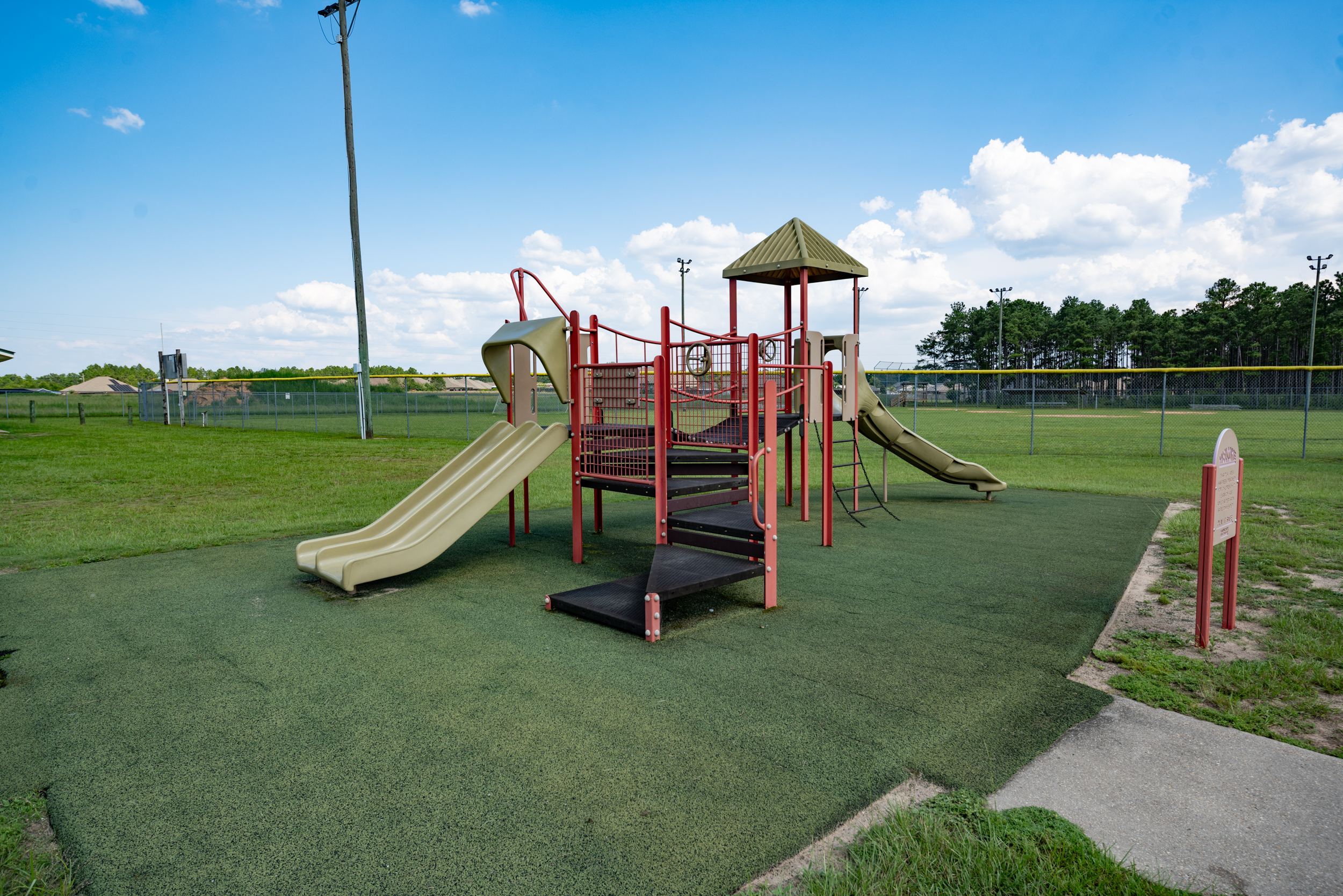 A playground with slides, climbing structures, and a green rubberized surface, surrounded by grass and fencing, with some trees visible in the background.