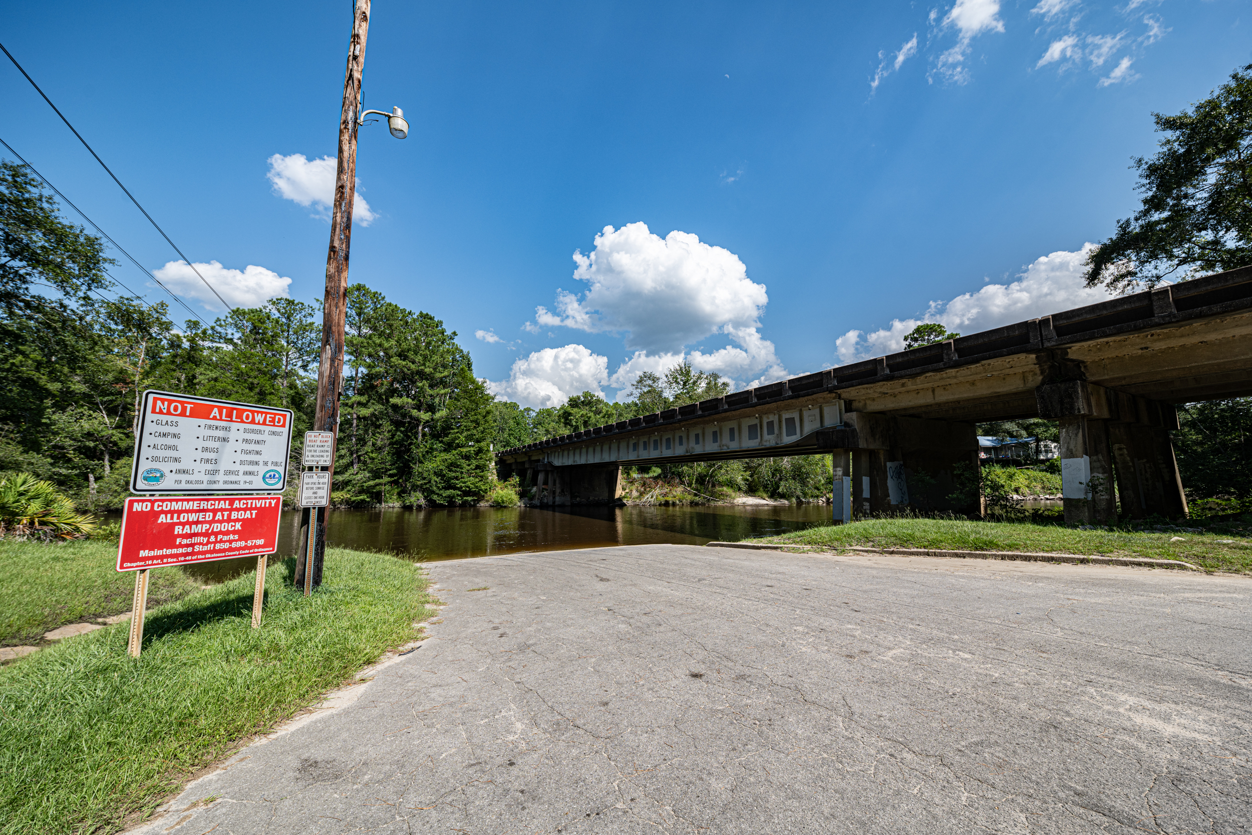 A paved road leads to a boat ramp next to a river, with a bridge overhead. A sign reads,