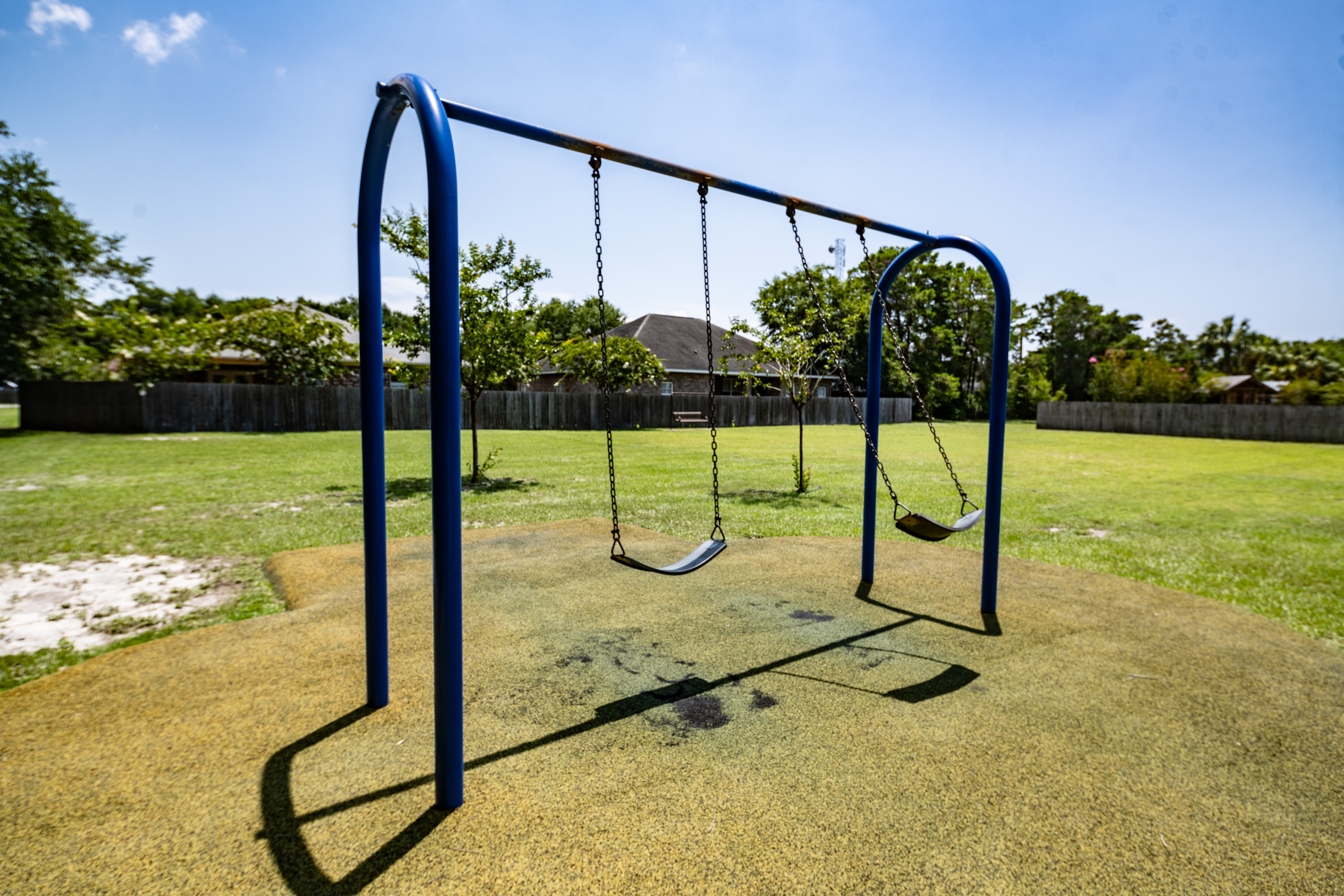A playground swing set with two empty swings under a clear blue sky, surrounded by grassy areas and trees in the background.