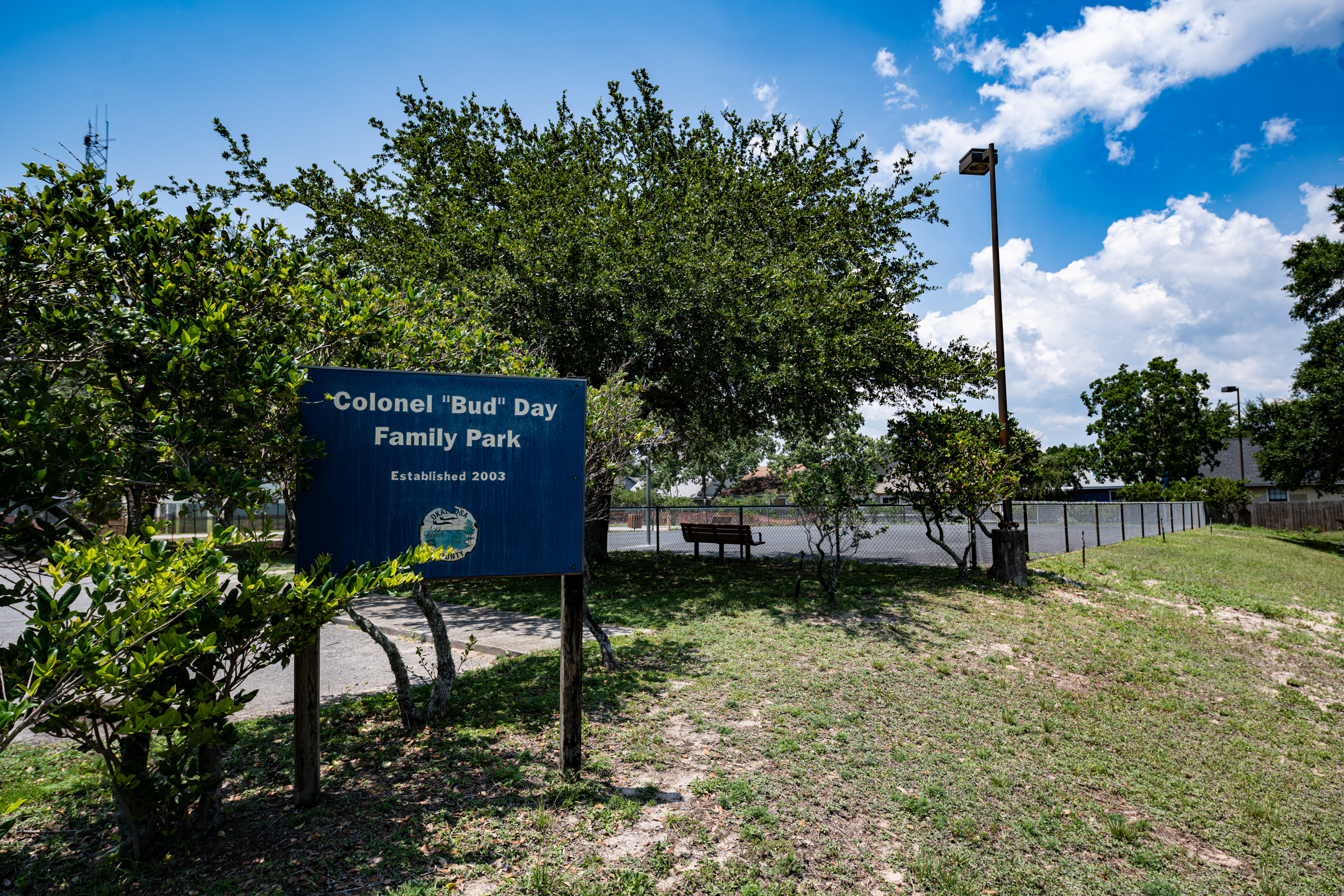 Sign reading “Colonel 'Bud' Day Family Park Established 2002” in front of trees and a paved area with a park bench. Cloudy sky and grassy foreground.