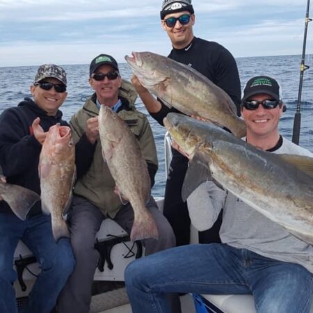 Five men on a boat holding large fish they caught, smiling at the camera. Fishing rods are visible in the background with the sea and cloudy sky behind them.