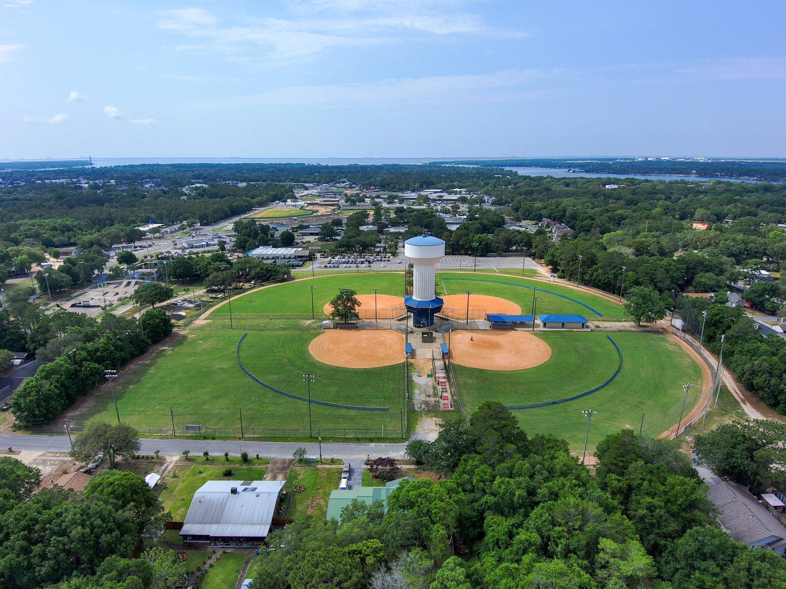 Aerial view of a baseball field with a central water tower, surrounded by trees and residential areas, under a clear sky.