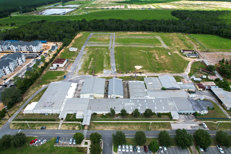 Aerial view of an industrial area with warehouses in the foreground, adjacent residential buildings, and expansive green fields in the background. Multiple vehicles are parked along the main road.