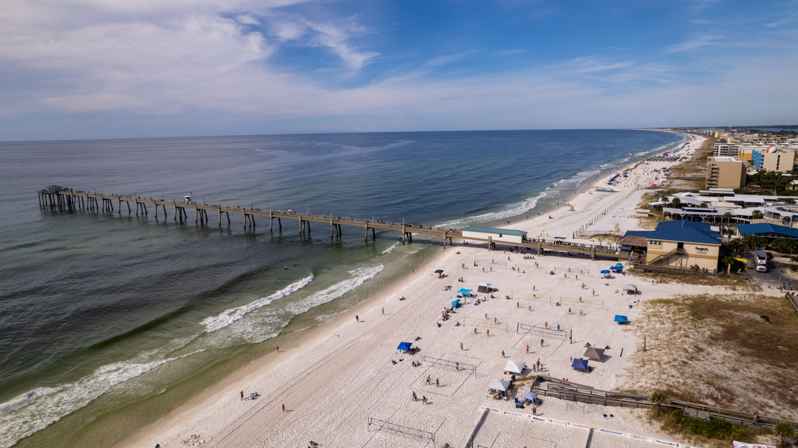 Aerial view of a long pier extending into the ocean with people on a sandy beach, buildings lining the shoreline, and clear blue skies above.
