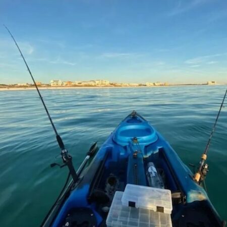 View from a blue kayak on calm waters with two fishing rods, facing towards a distant shoreline with buildings.