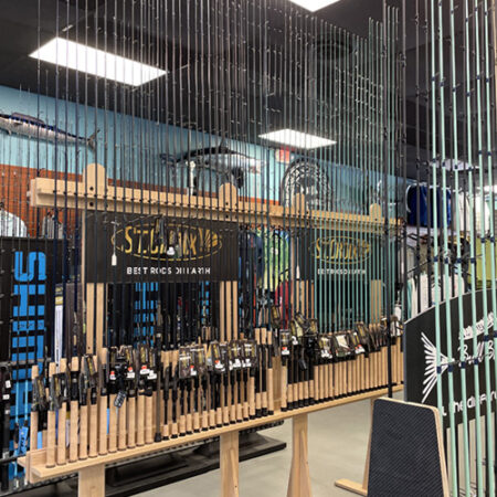 A fishing gear store display with numerous fishing rods of various brands on wooden racks. The background shows product logos and mounted fish decor on the wall.
