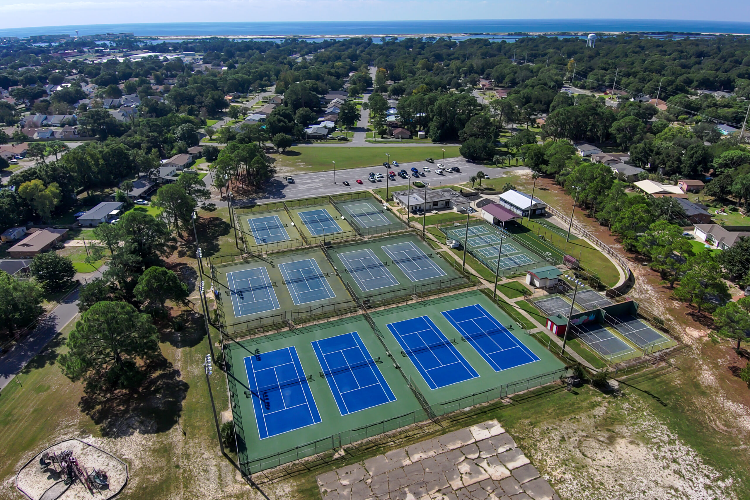 Aerial view of an outdoor tennis court complex surrounded by trees and residential buildings with a distant view of a large body of water on the horizon.