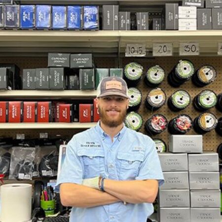 A person wearing a blue shirt and hat stands with folded arms in front of a store display of fishing gear, including rods, reels, and boxes of accessories.