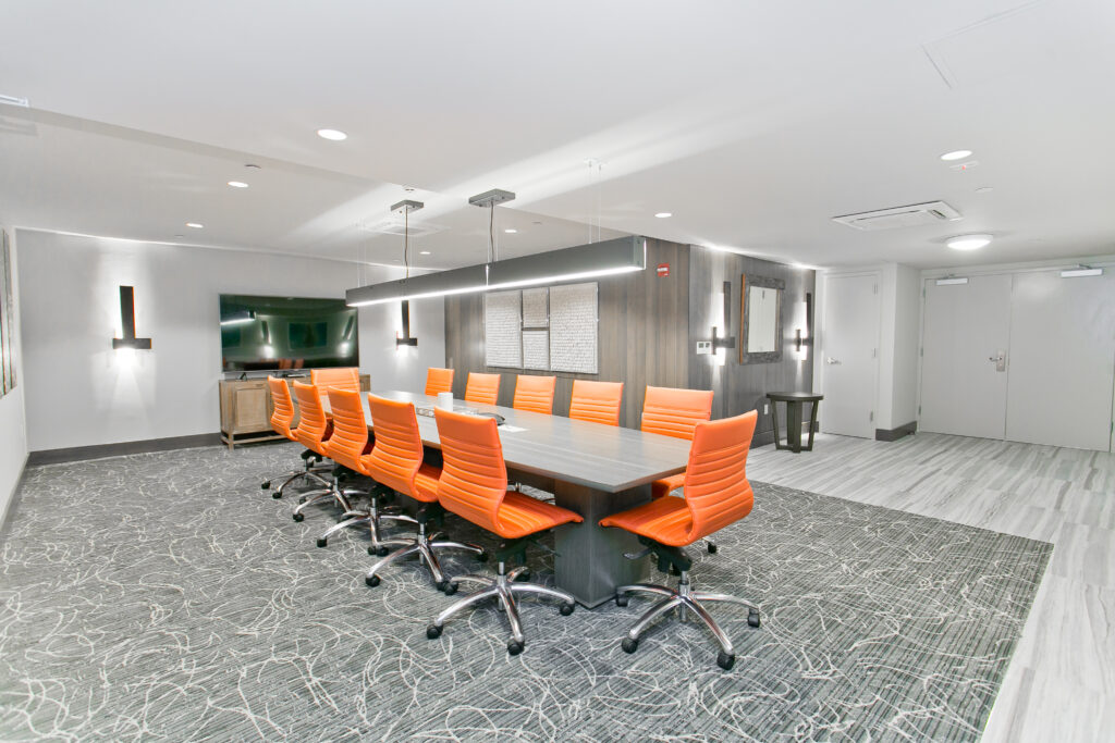 Modern conference room with a long table surrounded by ten orange chairs, a large TV, and contemporary lighting fixtures.