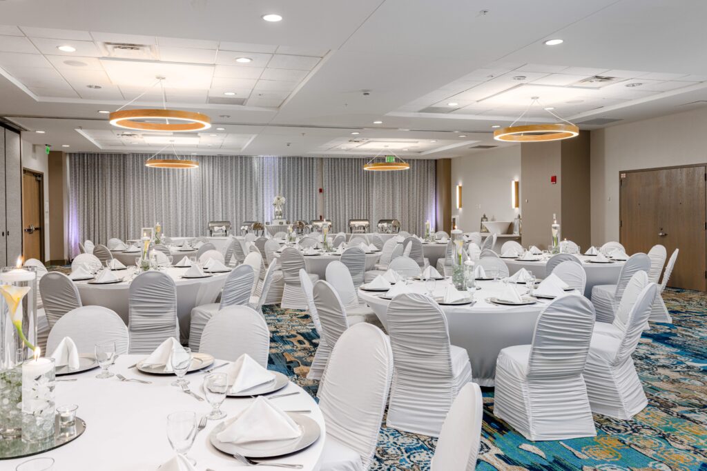 A banquet hall is set with round tables covered in white tablecloths and chairs draped in white covers. Each table has neatly arranged place settings and minimalist floral centerpieces.