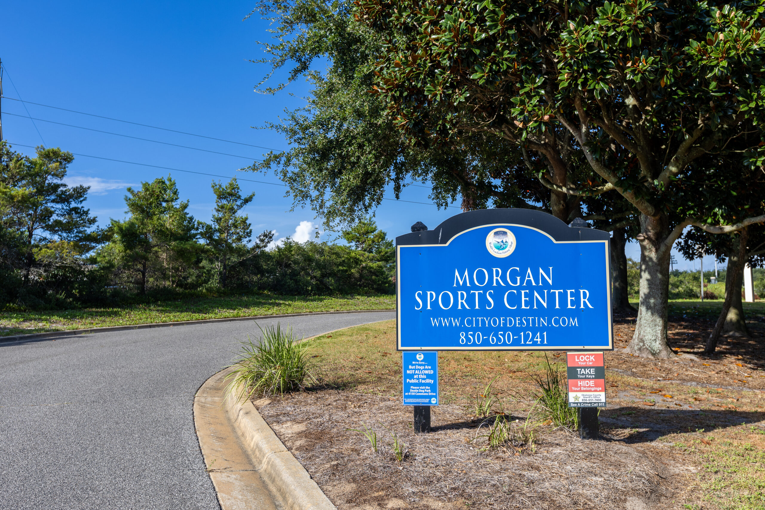A blue sign for Morgan Sports Center with contact information (website and phone number) stands beside a curved road and some trees.