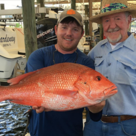 Two men are smiling at a dock while one holds a large red snapper fish. Boats and fishing gear are visible in the background.