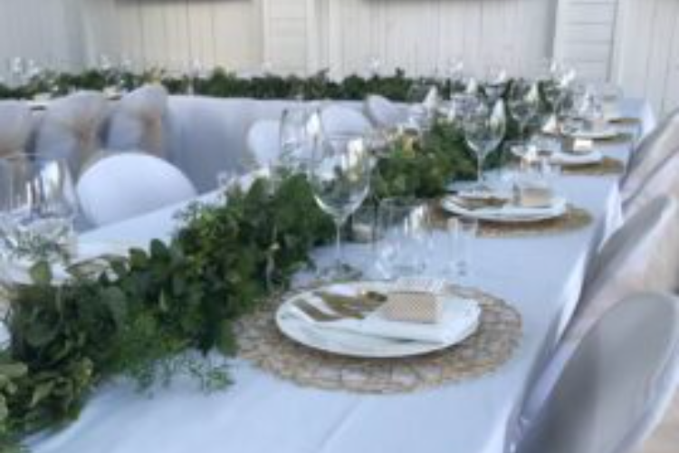 Elegant table setting with white tablecloths, green foliage runners, and place settings with white plates, glasses, and wicker placemats arranged for an event.