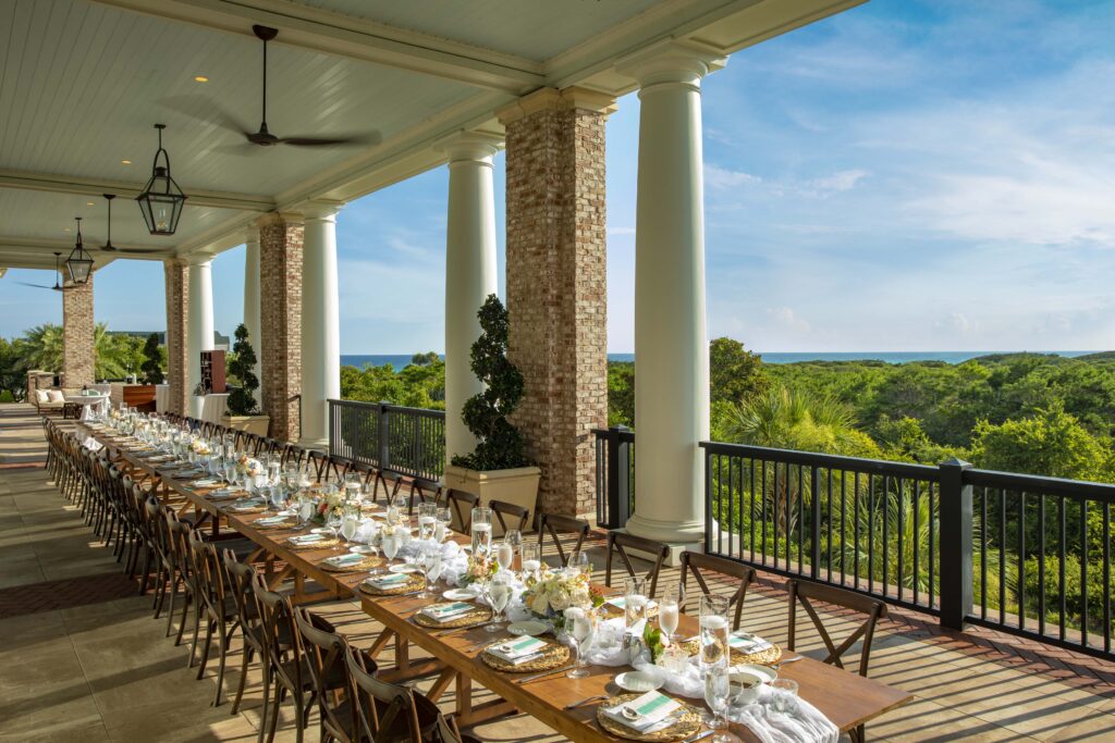 A long, wooden banquet table set for an outdoor meal on a covered veranda with a view of lush greenery and the ocean in the distance.