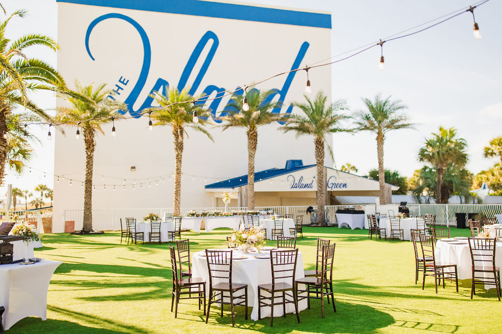 Outdoor event space with round tables and chairs set up on a grassy area. A white building with the word