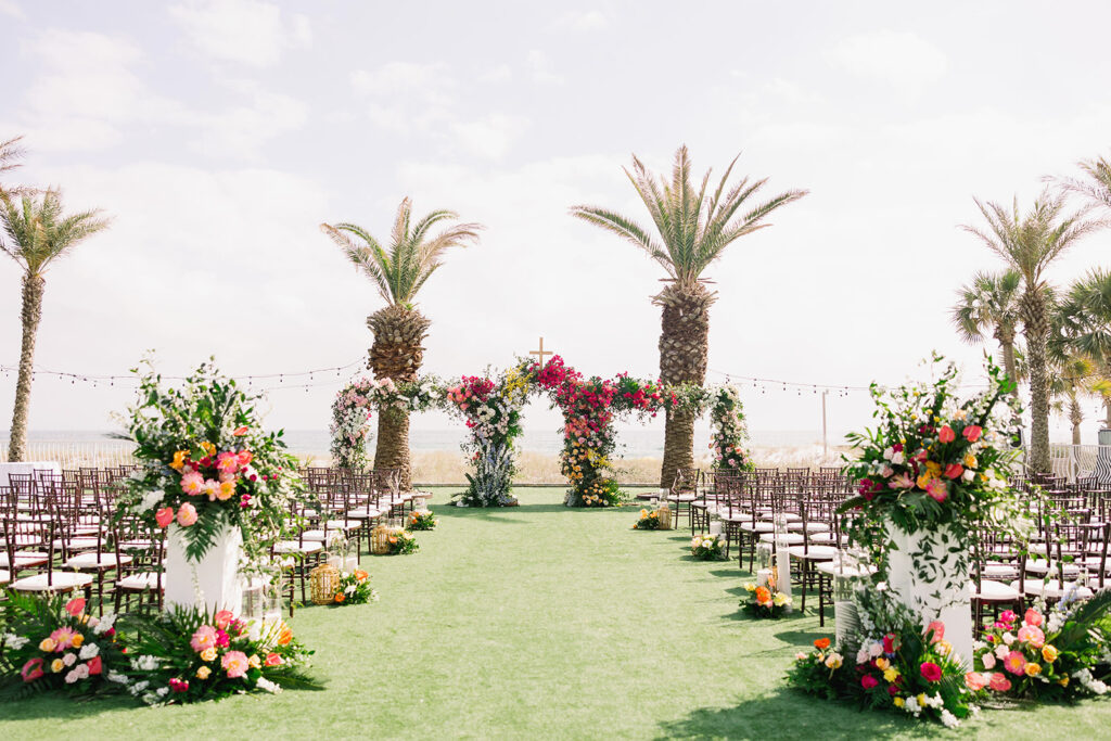 An outdoor wedding setup featuring two rows of chairs on either side of a grass aisle, leading to an archway decorated with colorful flowers and greenery, with palm trees and string lights in the background.