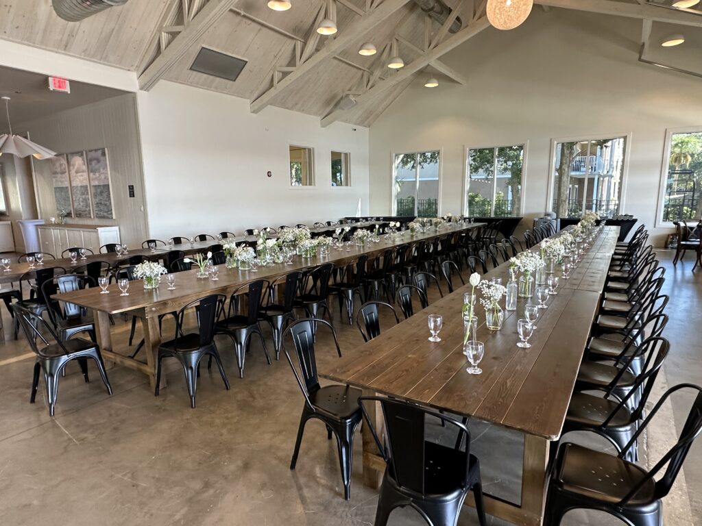 A long wooden table setup for a formal event with black chairs and floral centerpieces in a large room with vaulted ceilings and large windows.